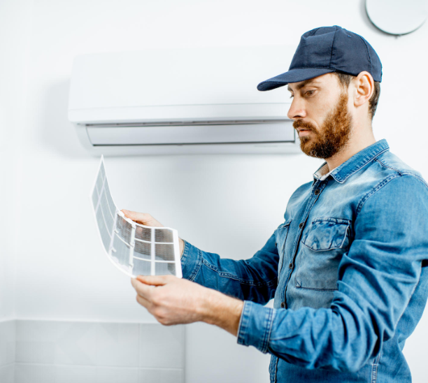A man wearing a blue cap and denim shirt inspects an air conditioner filter. He stands in a room with a white wall-mounted unit, carefully examining the filter against the plain background, showcasing his expertise in air con services near me.