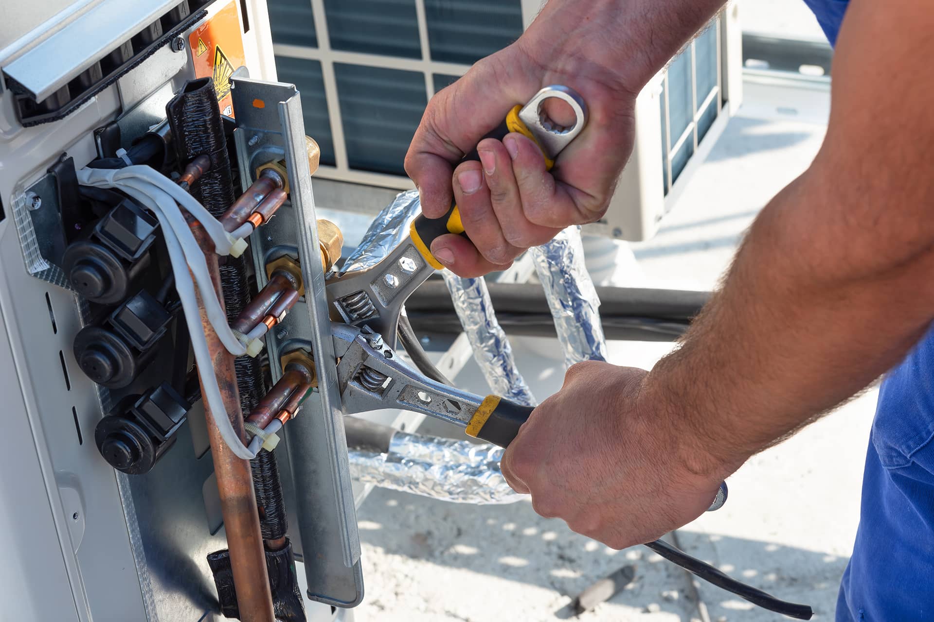Close-up of a person using pliers to adjust copper piping on an outdoor HVAC unit, likely on a rooftop. They wear blue clothing amidst insulated cables and tubes, expertly performing air conditioner services. The focus is on the hands and tools essential for this maintenance work.