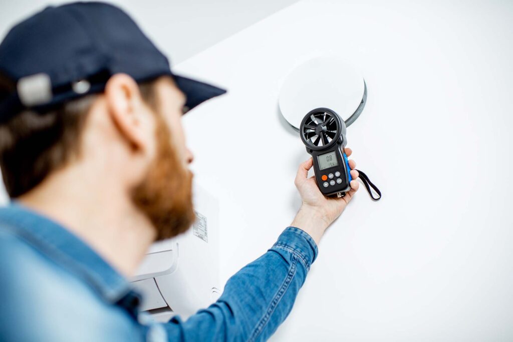 A man in a blue denim shirt and cap uses an anemometer to measure airflow from a round ceiling vent, showcasing the precision of air con services. His hand holds the device, displaying a digital reading, highlighting the focus on indoor measurement accuracy.