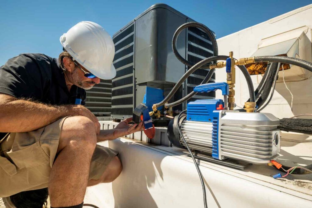 A worker in a white hard hat and sunglasses inspects HVAC equipment on a rooftop, offering specialized air conditioner services. He crouches beside a large unit, using tools and pressure gauges. The sky is clear and blue, and he wears a black shirt and tan shorts. A vacuum pump is visible in the foreground.