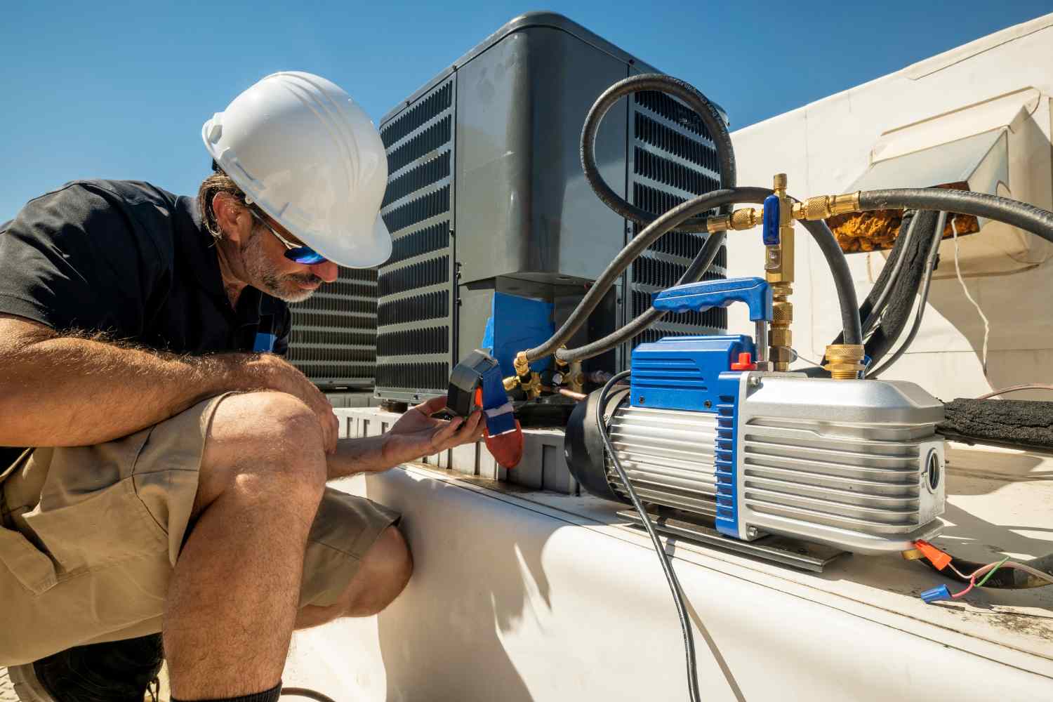 A worker in a white hard hat and sunglasses inspects HVAC equipment on a rooftop, offering specialized air conditioner services. He crouches beside a large unit, using tools and pressure gauges. The sky is clear and blue, and he wears a black shirt and tan shorts. A vacuum pump is visible in the foreground.
