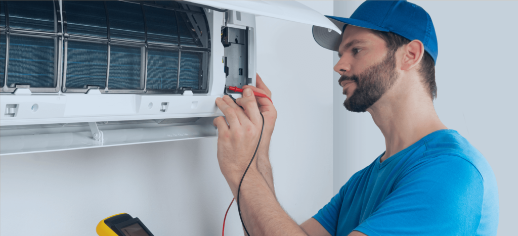 A man in a blue shirt and cap is meticulously repairing an air conditioning unit, using a multimeter with red and black probes to test the connections. The units cover is open, revealing its inner components. For reliable air con services near me, his skills underscore the expertise needed for quality maintenance work.