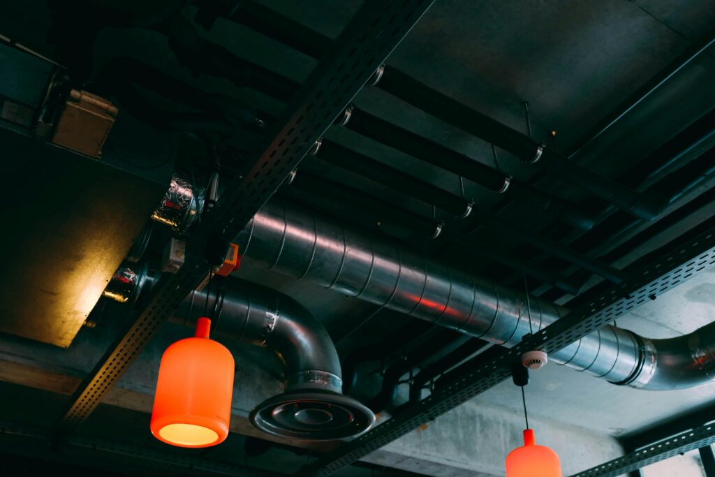 A view of an industrial ceiling showcases exposed ducts and pipes, with two orange pendant lights casting a warm glow. Metal beams and wiring enhance the aesthetic. Amidst the metallic, reflective ducts, youd imagine finding air con services near me to keep things cool and efficient.