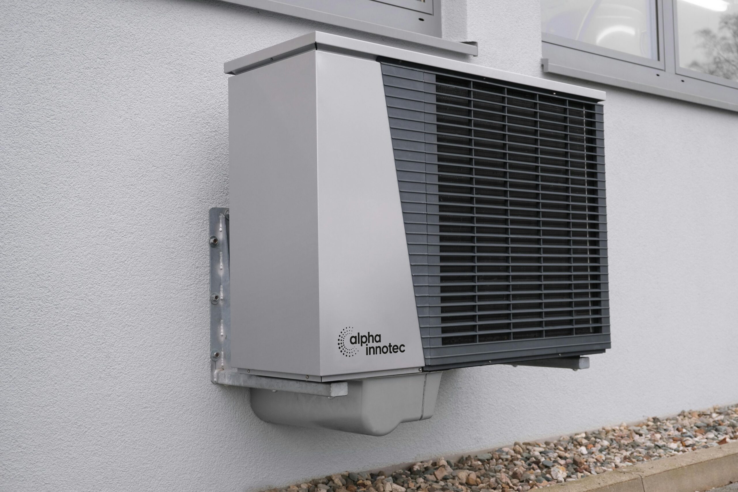 A sleek, rectangular metal heat pump unit with a grid-patterned cover, perfect for Miamis scorching days, is mounted on a white textured wall. The alpha innotec logo adorns the lower left corner. Below are small stones at the walls base, and a window is partially visible above.