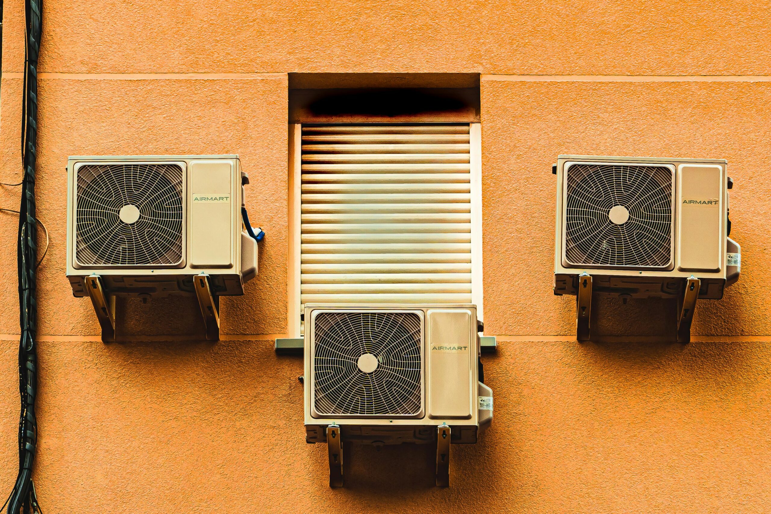 Three air conditioning units are mounted on an orange stucco wall, surrounding a central window with closed horizontal shutters. Each unit has a fan visible through a metal grill, evenly spaced for symmetry. A black cable runs vertically on the left side, indicating professional air con services attendance.