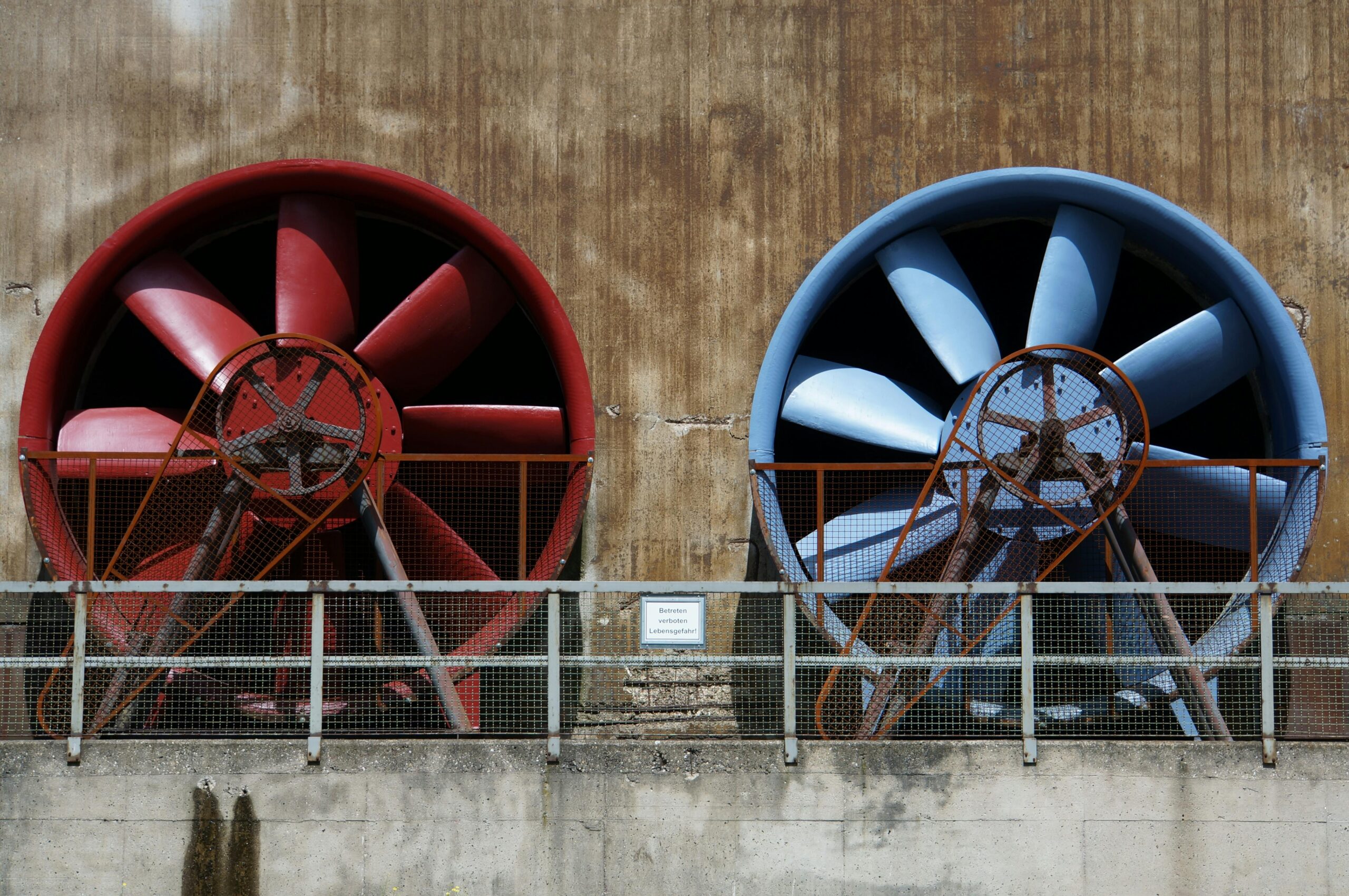 Two large industrial ventilation fans, reminiscent of robust air con services, are installed side by side on a weathered concrete wall. The left is red, the right blue, both shielded by metal grilles and rusted frames, with a small sign between them hinting at air conditioner unit replacement costs.
