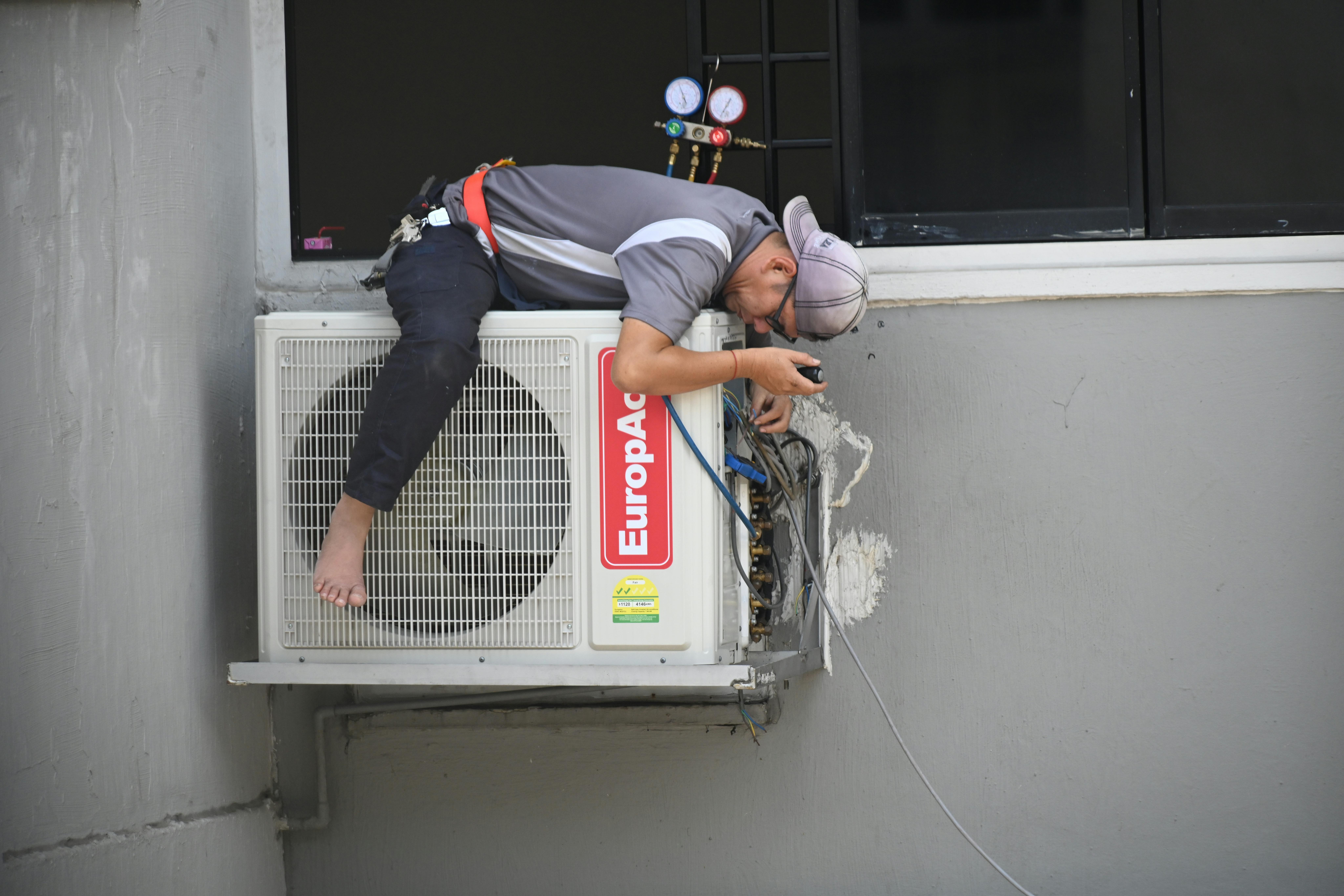 A man in a gray and black uniform is diligently working on an air conditioning unit in Miami. Lying atop the unit, he checks connections with a gauge, feet dangling over the side of the gray building with a window nearby.