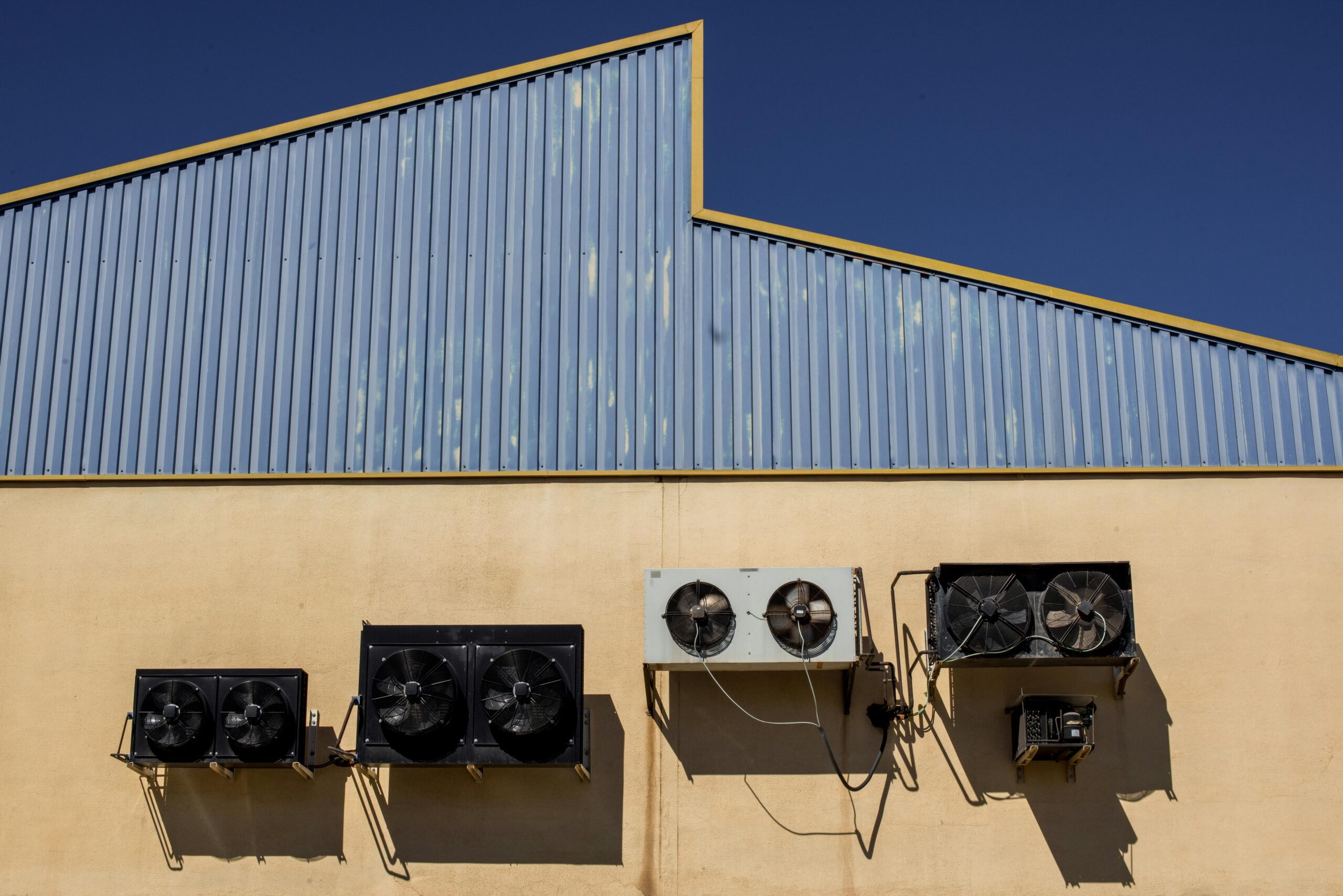 Exterior view of a beige building featuring five air conditioning units mounted on the wall. Above, a corrugated metal roof in blue angles sharply against Miamis clear, deep blue sky. The units vary in size and are connected by visible cables. Shadows accentuate the structure’s geometry.