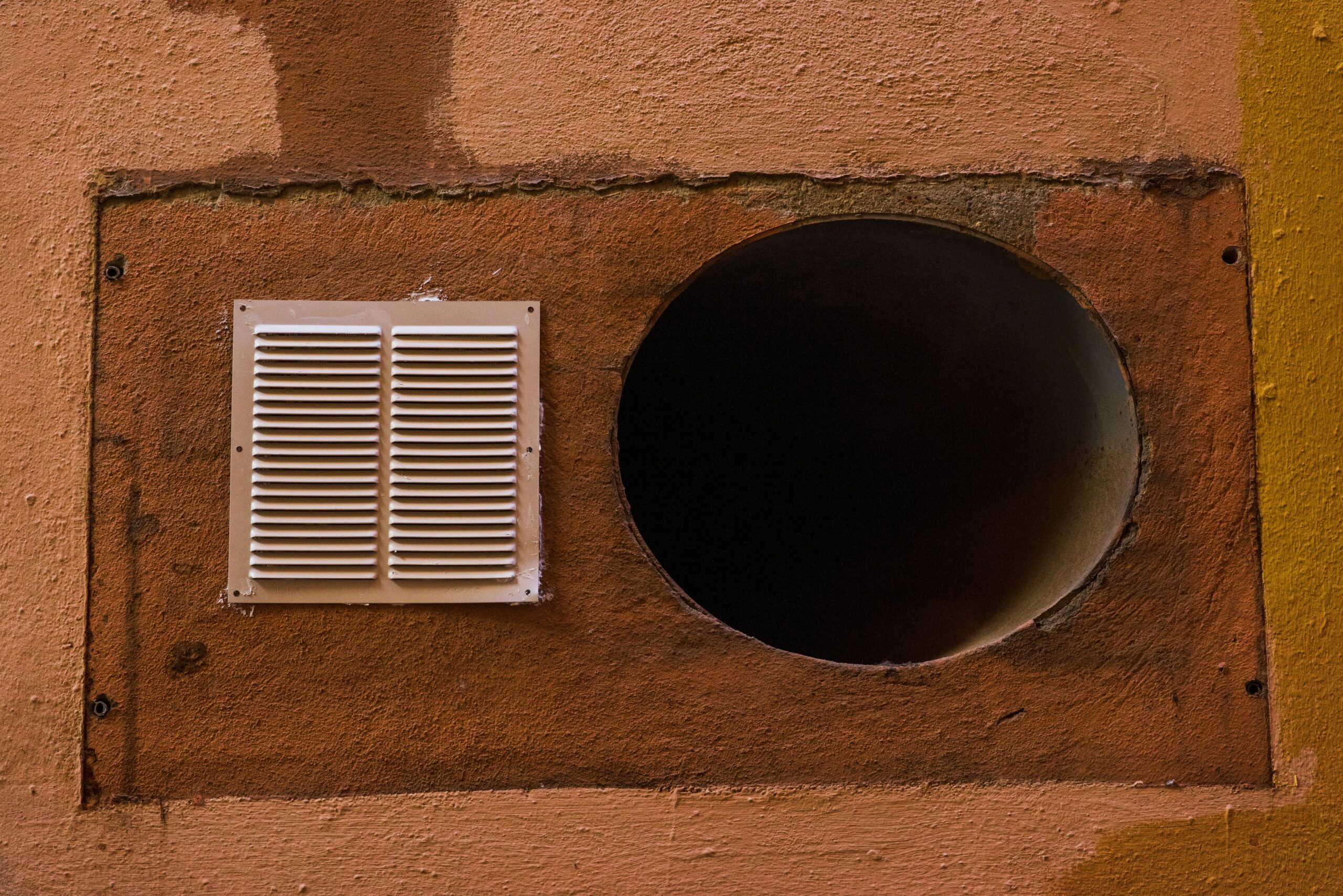 A textured wall with two ventilation openings—perfect for those considering air conditioner Miami installations. The left has a white rectangular vent cover, the right a large, dark circular hole. Painted in shades of brown and yellow, its marked by visible patches and wear.